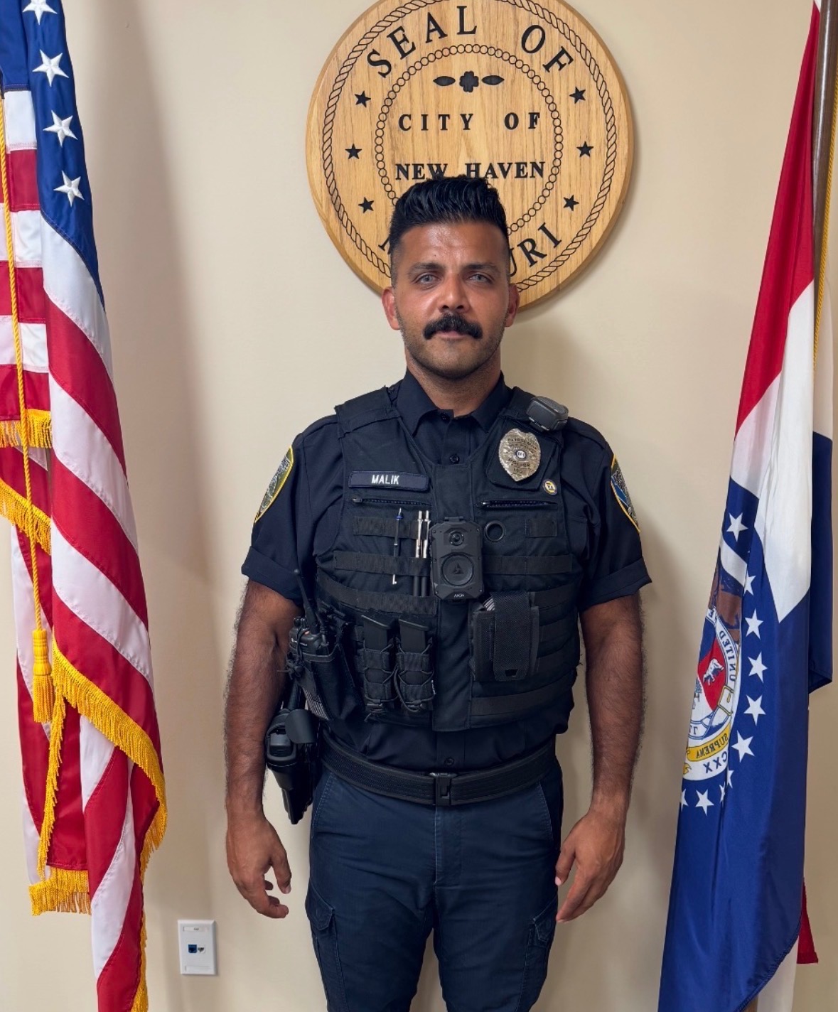 Officer in black tactical uniform stands before “Seal of the City of New Haven” with U.S. and Connecticut flags.